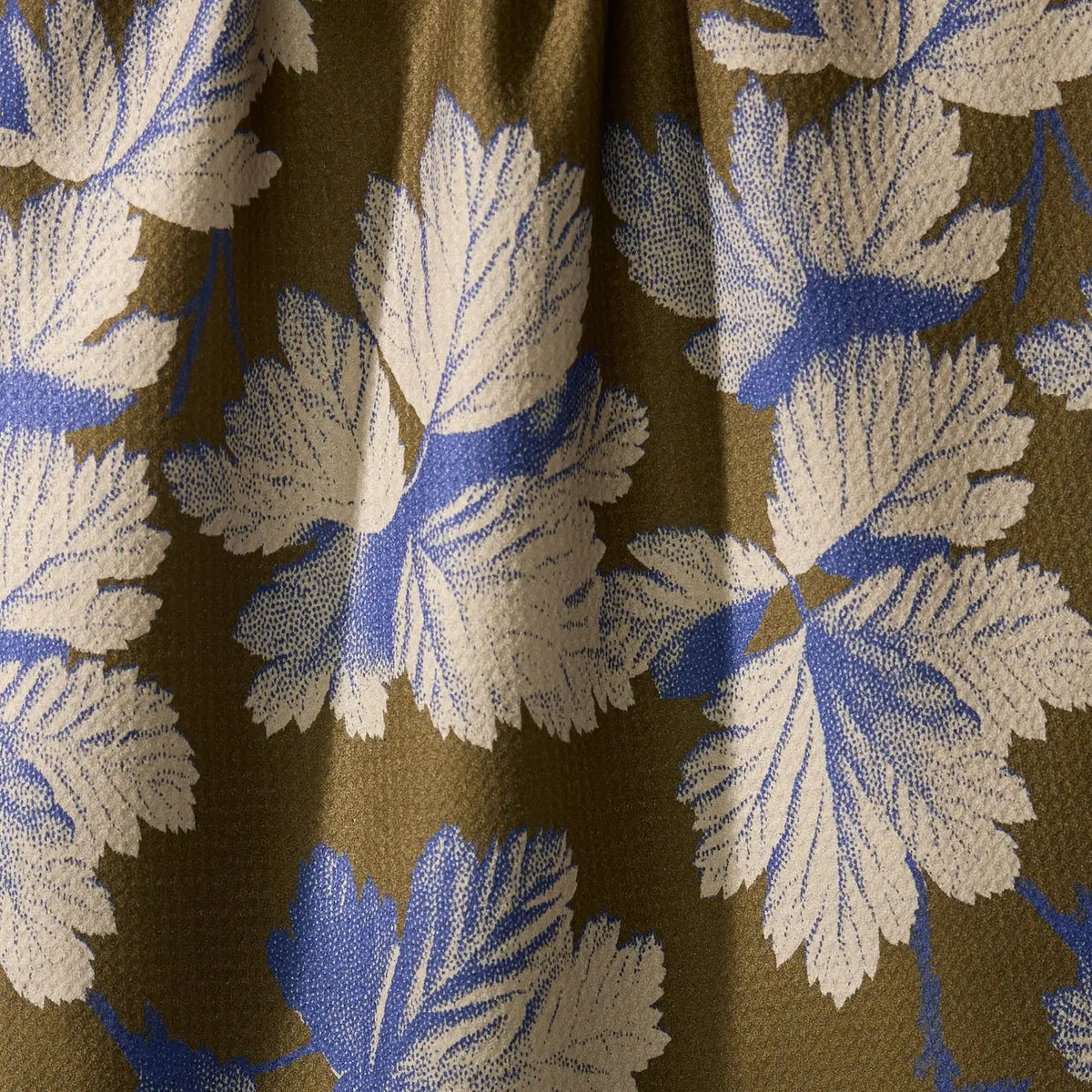 A close-up of a fabric or wallpaper pattern featuring large cream-colored leaf motifs with blue centers on a warm olive-brown background.